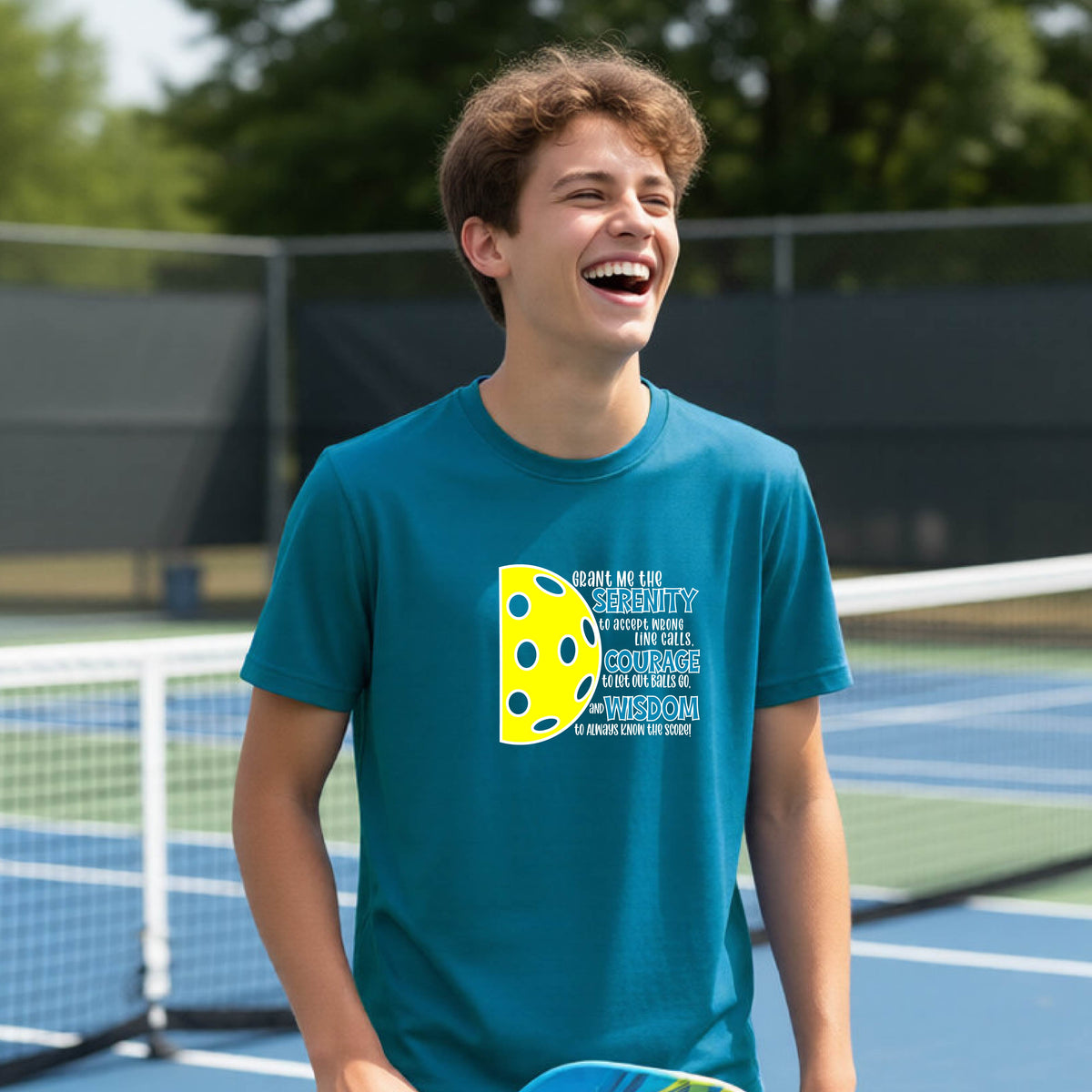A young person on a pickleball court wearing a teal short-sleeve t-shirt with a graphic of the Serenity Prayer made for pickleball:  “Grant me the serenity to Accept Wrong Line Calls; Courage to Let Out Balls Go; and, Wisdom to Always Know the Score” next to a half-pickleball icon, standing on a pickleball court.