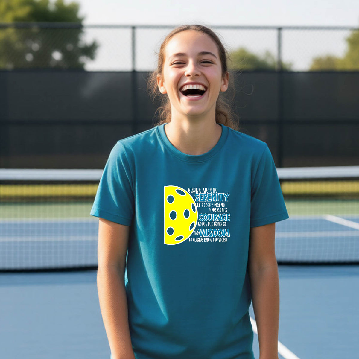A young person on a pickleball court wearing a teal short-sleeve t-shirt with a graphic of the Serenity Prayer made for pickleball:  “Grant me the serenity to Accept Wrong Line Calls; Courage to Let Out Balls Go; and, Wisdom to Always Know the Score” next to a half-pickleball icon, standing on a pickleball court.