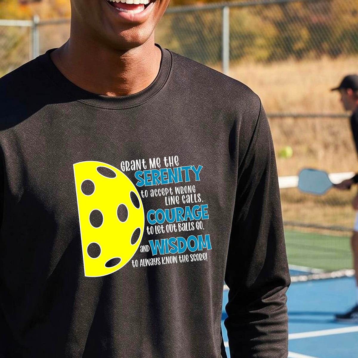 A man on a pickleball court wearing a black long-sleeve t-shirt with a graphic of the Serenity Prayer made for pickleball:  “Grant me the serenity to Accept Wrong Line Calls; Courage to Let Out Balls Go; and, Wisdom to Always Know the Score” next to a half-pickleball icon, standing on a pickleball court.