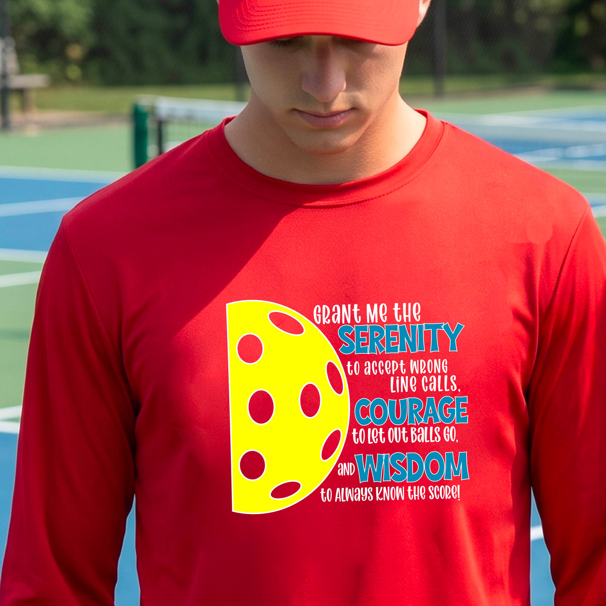 A man on a pickleball court wearing a red long-sleeve t-shirt with a graphic of the Serenity Prayer made for pickleball:  “Grant me the serenity to Accept Wrong Line Calls; Courage to Let Out Balls Go; and, Wisdom to Always Know the Score” next to a half-pickleball icon, standing on a pickleball court.