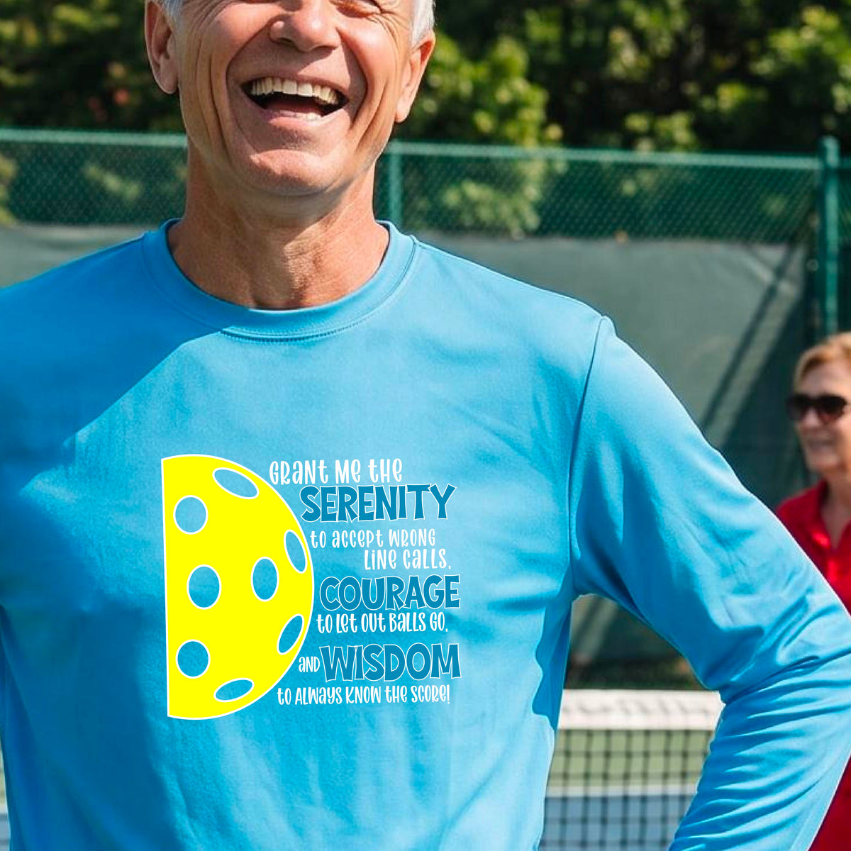 A man on a pickleball court wearing a bright blue long-sleeve t-shirt with a graphic of the Serenity Prayer made for pickleball:  “Grant me the serenity to Accept Wrong Line Calls; Courage to Let Out Balls Go; and, Wisdom to Always Know the Score” next to a half-pickleball icon, standing on a pickleball court.