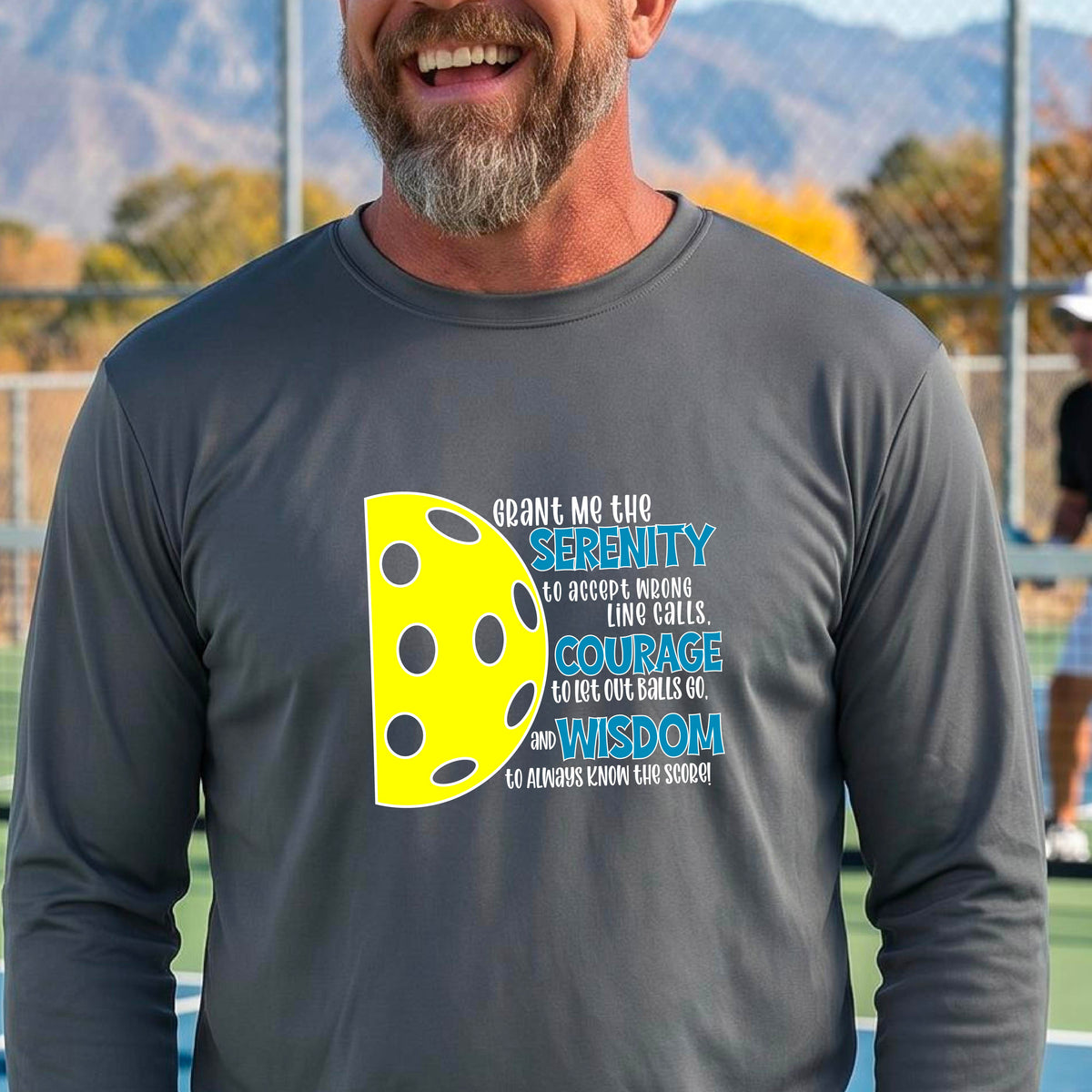 A man on a pickleball court wearing a gray long-sleeve t-shirt with a graphic of the Serenity Prayer made for pickleball:  “Grant me the serenity to Accept Wrong Line Calls; Courage to Let Out Balls Go; and, Wisdom to Always Know the Score” next to a half-pickleball icon, standing on a pickleball court.