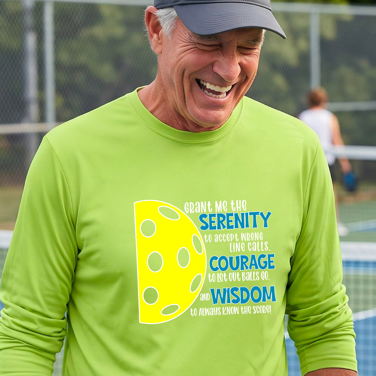 A man on a pickleball court wearing a lime green long-sleeve t-shirt with a graphic of the Serenity Prayer made for pickleball:  “Grant me the serenity to Accept Wrong Line Calls; Courage to Let Out Balls Go; and, Wisdom to Always Know the Score” next to a half-pickleball icon, standing on a pickleball court.