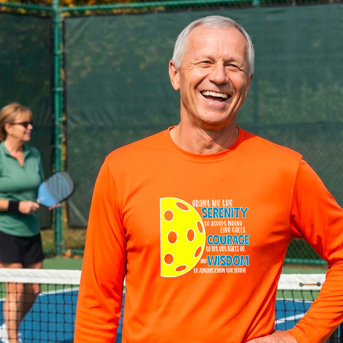A man on a pickleball court wearing an orange long-sleeve t-shirt with a graphic of the Serenity Prayer made for pickleball:  “Grant me the serenity to Accept Wrong Line Calls; Courage to Let Out Balls Go; and, Wisdom to Always Know the Score” next to a half-pickleball icon, standing on a pickleball court.