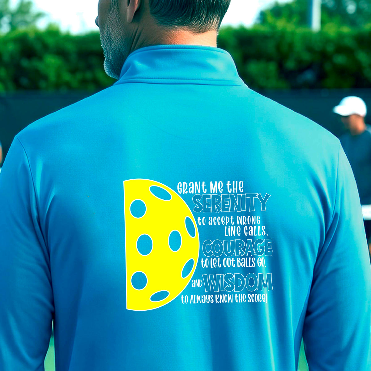 A man on a pickleball court facing backwards, wearing a bright blue quarter-zip shirt with a graphic of the Serenity Prayer made for pickleball:  “Grant me the serenity to Accept Wrong Line Calls; Courage to Let Out Balls Go; and, Wisdom to Always Know the Score” next to a half-pickleball icon, standing on a pickleball court.