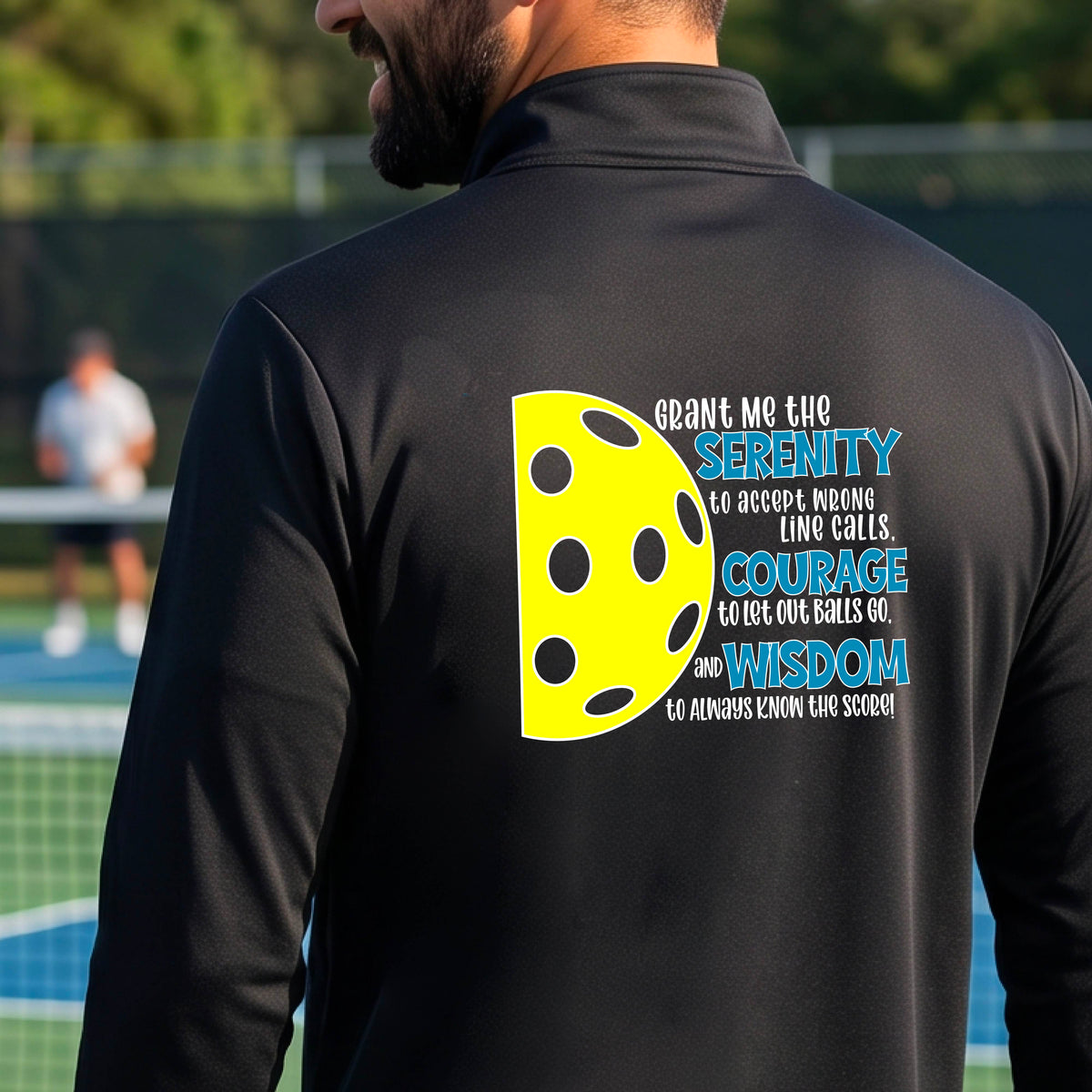A man on a pickleball court facing backwards, wearing a black quarter-zip shirt with a graphic of the Serenity Prayer made for pickleball:  “Grant me the serenity to Accept Wrong Line Calls; Courage to Let Out Balls Go; and, Wisdom to Always Know the Score” next to a half-pickleball icon, standing on a pickleball court.