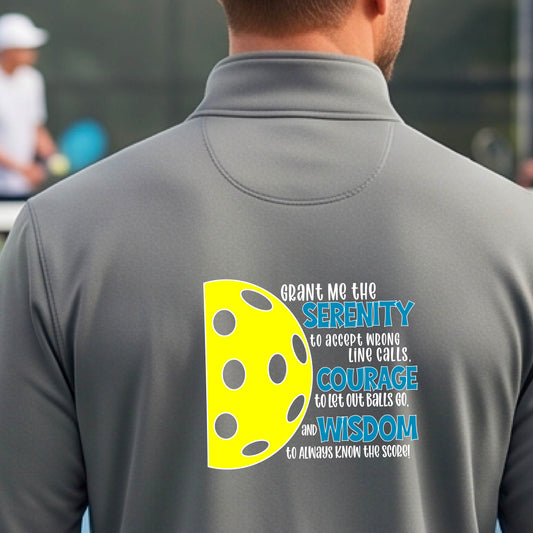 A man on a pickleball court facing backwards, wearing a gray quarter-zip shirt with a graphic of the Serenity Prayer made for pickleball:  “Grant me the serenity to Accept Wrong Line Calls; Courage to Let Out Balls Go; and, Wisdom to Always Know the Score” next to a half-pickleball icon, standing on a pickleball court.