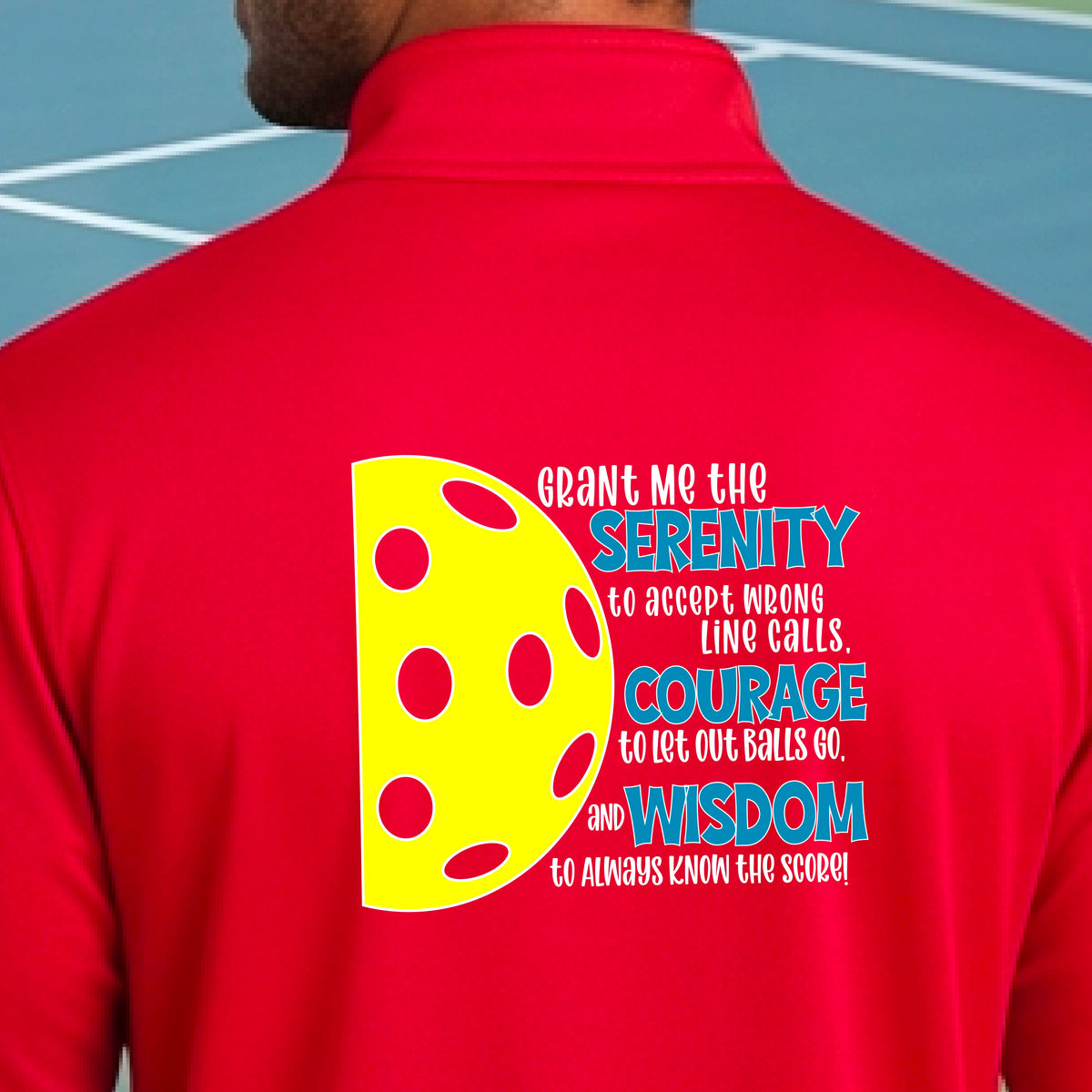 A man on a pickleball court facing backwards, wearing a red quarter-zip shirt with a graphic of the Serenity Prayer made for pickleball:  “Grant me the serenity to Accept Wrong Line Calls; Courage to Let Out Balls Go; and, Wisdom to Always Know the Score” next to a half-pickleball icon, standing on a pickleball court.