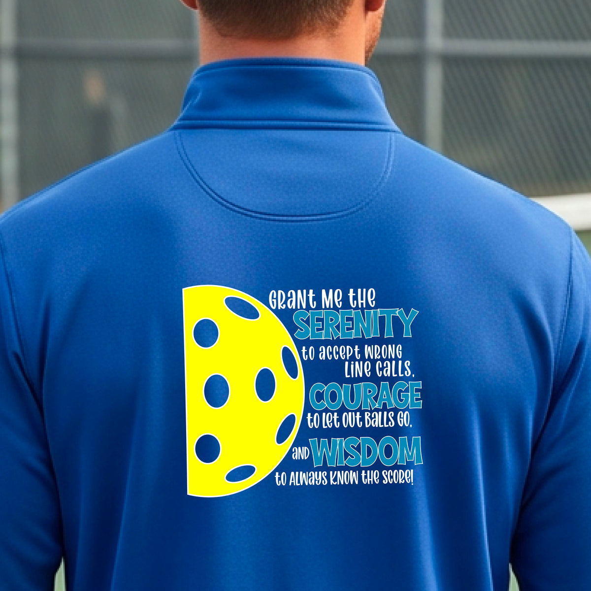 A man on a pickleball court facing backwards, wearing a royal blue quarter-zip shirt with a graphic of the Serenity Prayer made for pickleball:  “Grant me the serenity to Accept Wrong Line Calls; Courage to Let Out Balls Go; and, Wisdom to Always Know the Score” next to a half-pickleball icon, standing on a pickleball court.