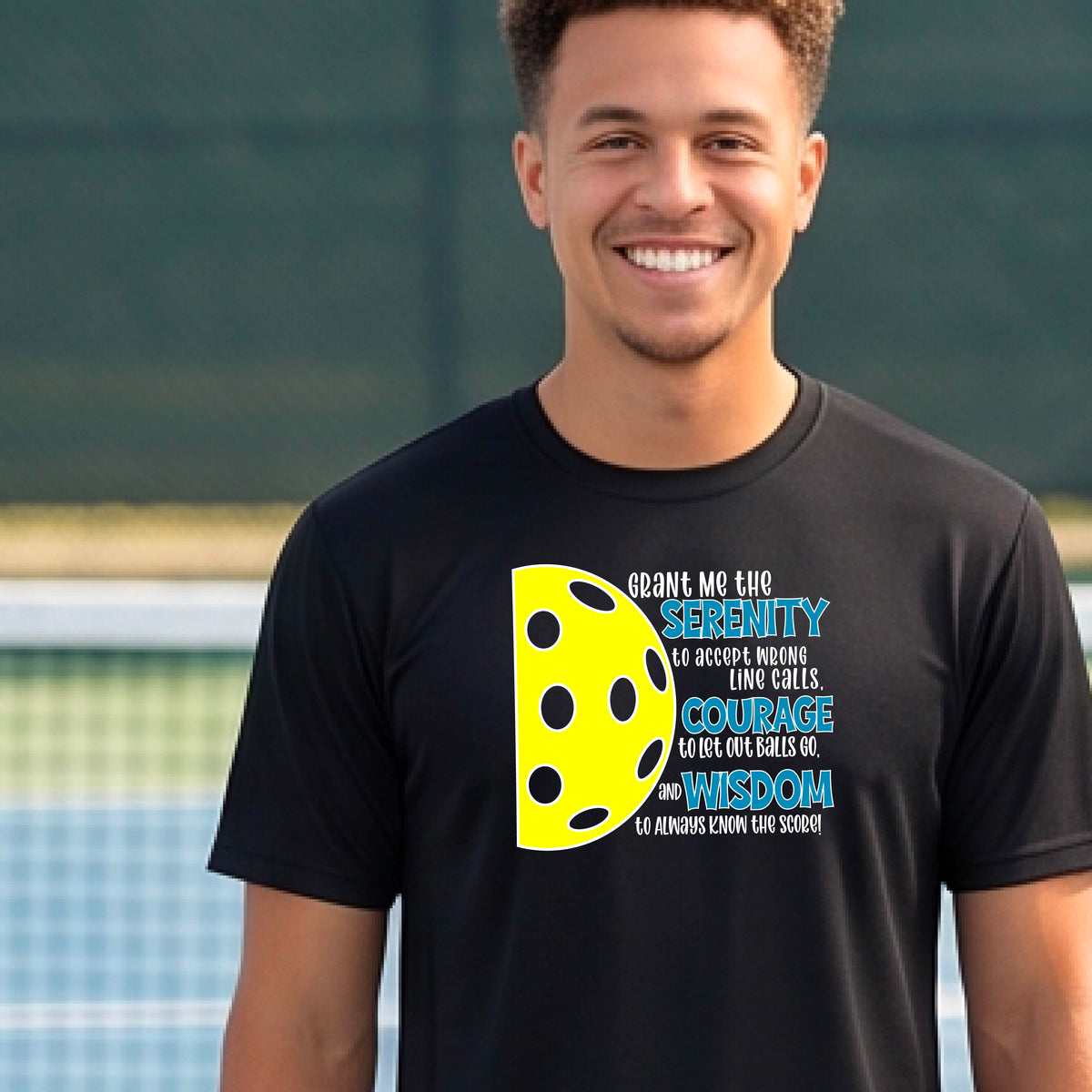 A man on a pickleball court wearing a black long-sleeve t-shirt with a graphic of the Serenity Prayer made for pickleball:  “Grant me the serenity to Accept Wrong Line Calls; Courage to Let Out Balls Go; and, Wisdom to Always Know the Score” next to a half-pickleball icon, standing on a pickleball court.
