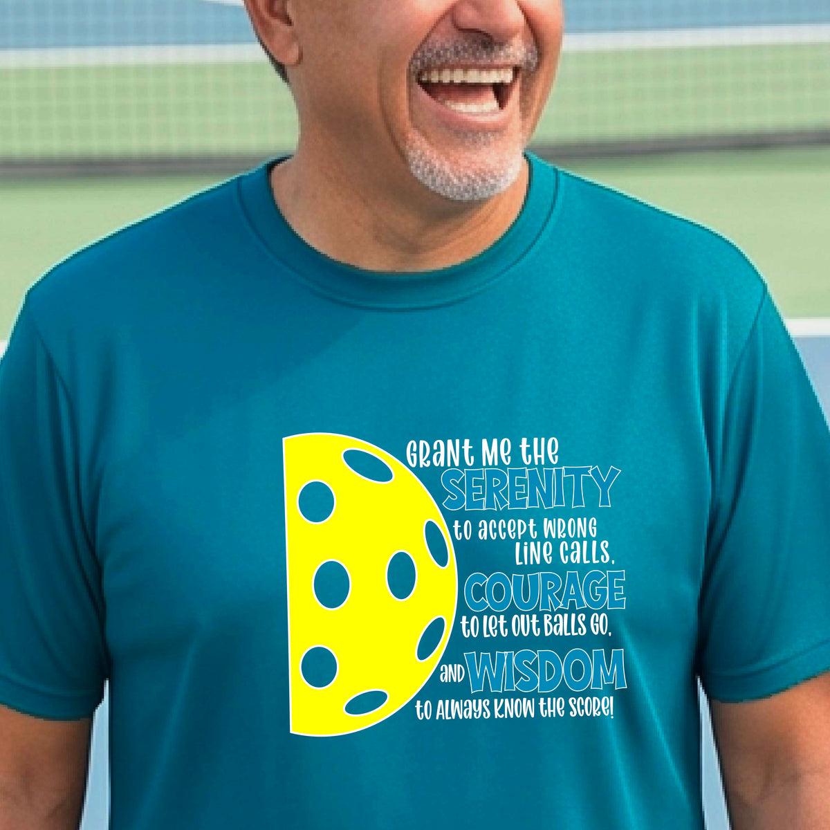A man on a pickleball court wearing a teal long-sleeve t-shirt with a graphic of the Serenity Prayer made for pickleball:  “Grant me the serenity to Accept Wrong Line Calls; Courage to Let Out Balls Go; and, Wisdom to Always Know the Score” next to a half-pickleball icon, standing on a pickleball court.