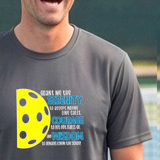 A man on a pickleball court wearing a dark gray long-sleeve t-shirt with a graphic of the Serenity Prayer made for pickleball:  “Grant me the serenity to Accept Wrong Line Calls; Courage to Let Out Balls Go; and, Wisdom to Always Know the Score” next to a half-pickleball icon, standing on a pickleball court.