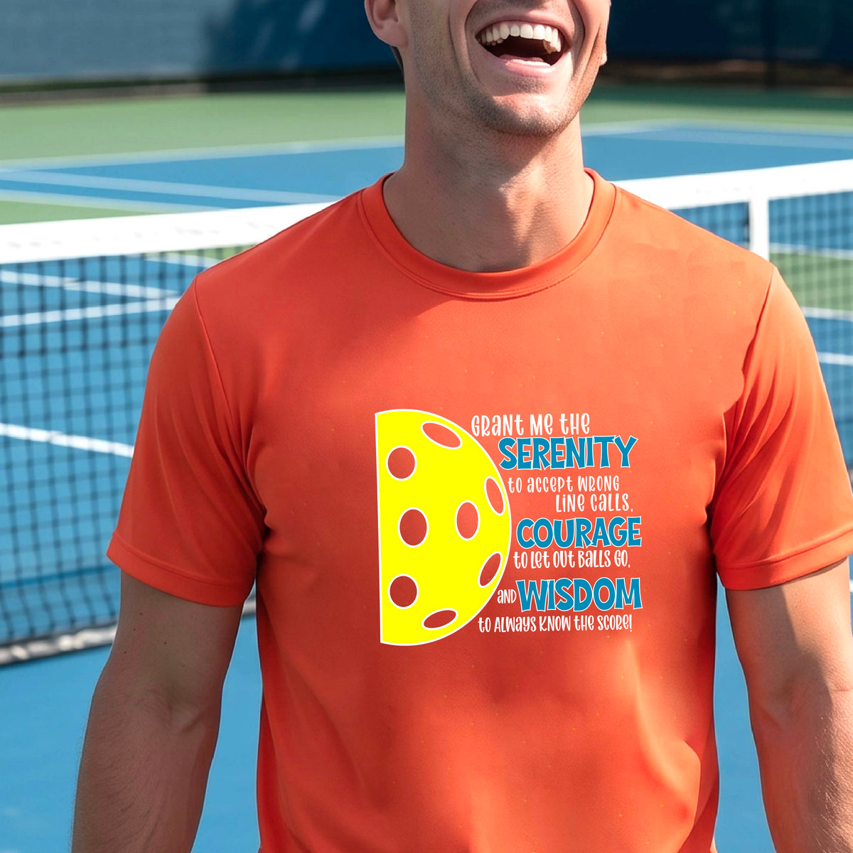 A man on a pickleball court wearing a orange long-sleeve t-shirt with a graphic of the Serenity Prayer made for pickleball:  “Grant me the serenity to Accept Wrong Line Calls; Courage to Let Out Balls Go; and, Wisdom to Always Know the Score” next to a half-pickleball icon, standing on a pickleball court.