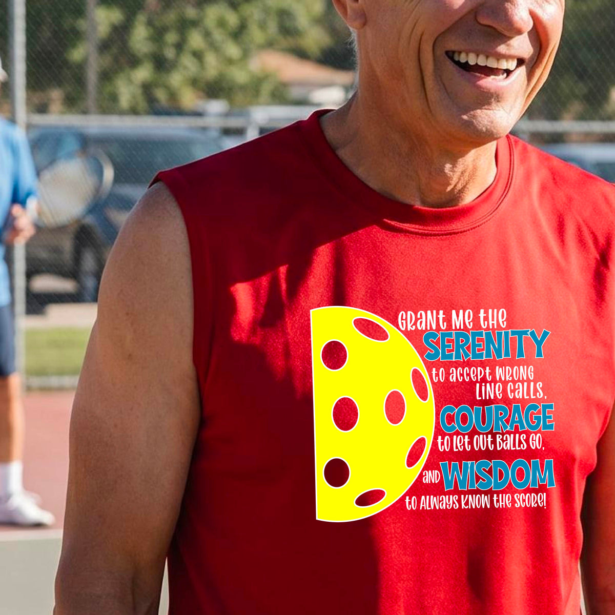 A man on a pickleball court wearing a red sleeveless t-shirt with a graphic of the Serenity Prayer made for pickleball:  “Grant me the serenity to Accept Wrong Line Calls; Courage to Let Out Balls Go; and, Wisdom to Always Know the Score” next to a half-pickleball icon, standing on a pickleball court.