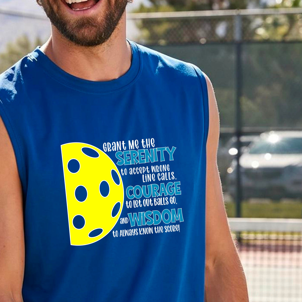 A man on a pickleball court wearing a royal blue sleeveless t-shirt with a graphic of the Serenity Prayer made for pickleball:  “Grant me the serenity to Accept Wrong Line Calls; Courage to Let Out Balls Go; and, Wisdom to Always Know the Score” next to a half-pickleball icon, standing on a pickleball court.