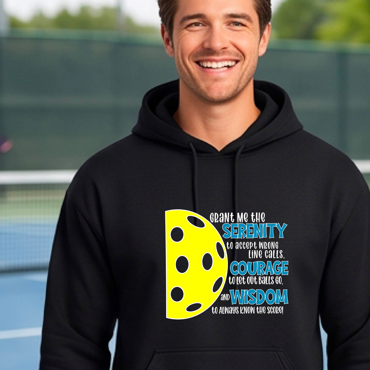 A man on a pickleball court wearing a black unisex hoodie pullover with a graphic of the Serenity Prayer made for pickleball:  “Grant me the serenity to Accept Wrong Line Calls; Courage to Let Out Balls Go; and, Wisdom to Always Know the Score” next to a half-pickleball icon, standing on a pickleball court.