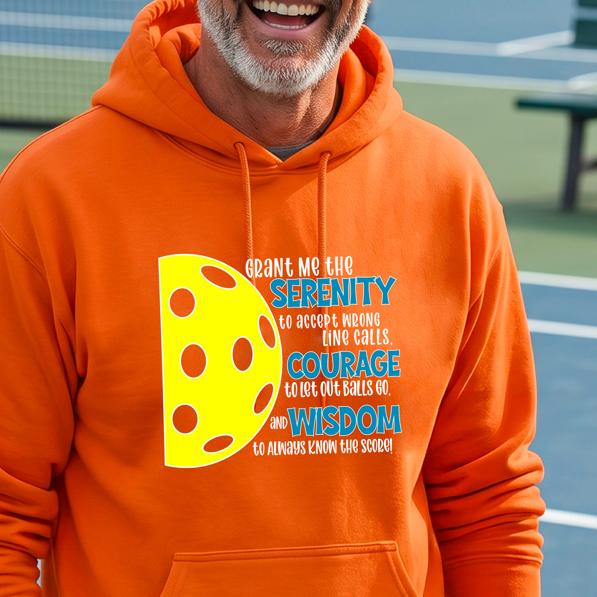 A man on a pickleball court wearing an orange unisex hoodie pullover with a graphic of the Serenity Prayer made for pickleball:  “Grant me the serenity to Accept Wrong Line Calls; Courage to Let Out Balls Go; and, Wisdom to Always Know the Score” next to a half-pickleball icon, standing on a pickleball court.