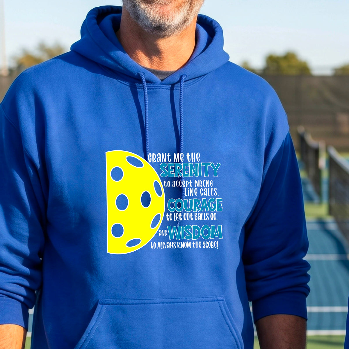 A man on a pickleball court wearing a royal blue unisex hoodie pullover with a graphic of the Serenity Prayer made for pickleball:  “Grant me the serenity to Accept Wrong Line Calls; Courage to Let Out Balls Go; and, Wisdom to Always Know the Score” next to a half-pickleball icon, standing on a pickleball court.