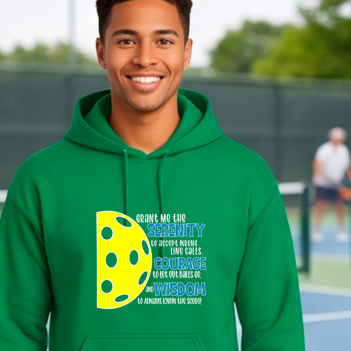 A man on a pickleball court wearing a green unisex hoodie pullover with a graphic of the Serenity Prayer made for pickleball:  “Grant me the serenity to Accept Wrong Line Calls; Courage to Let Out Balls Go; and, Wisdom to Always Know the Score” next to a half-pickleball icon, standing on a pickleball court.