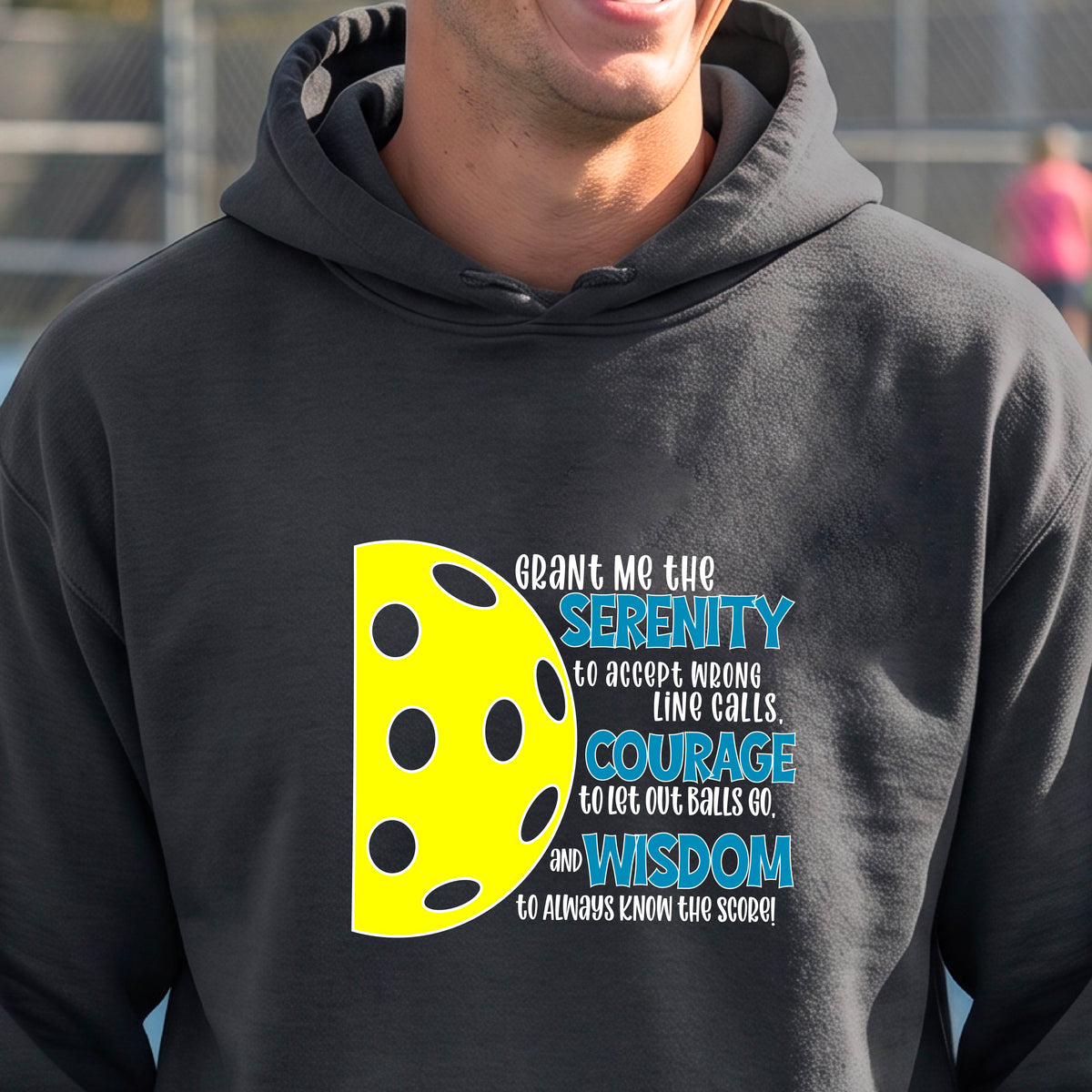 A man on a pickleball court wearing a gray unisex hoodie pullover with a graphic of the Serenity Prayer made for pickleball:  “Grant me the serenity to Accept Wrong Line Calls; Courage to Let Out Balls Go; and, Wisdom to Always Know the Score” next to a half-pickleball icon, standing on a pickleball court.