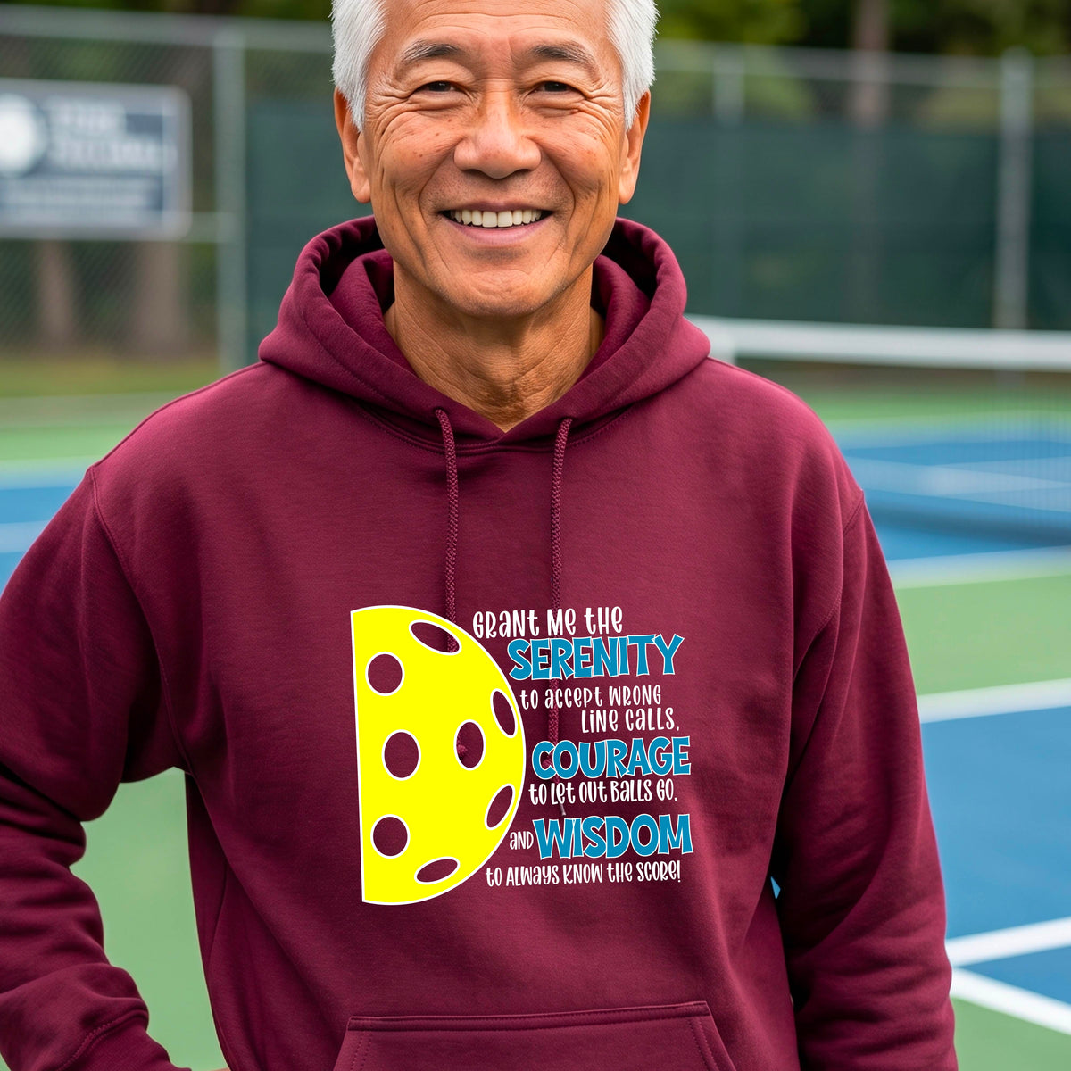 A man on a pickleball court wearing a maroon unisex hoodie pullover with a graphic of the Serenity Prayer made for pickleball:  “Grant me the serenity to Accept Wrong Line Calls; Courage to Let Out Balls Go; and, Wisdom to Always Know the Score” next to a half-pickleball icon, standing on a pickleball court.