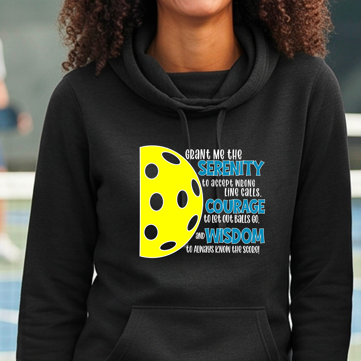 A woman on a pickleball court wearing a black cowl-neck hoodie pullover with a graphic of the Serenity Prayer made for pickleball:  “Grant me the serenity to Accept Wrong Line Calls; Courage to Let Out Balls Go; and, Wisdom to Always Know the Score” next to a half-pickleball icon, standing on a pickleball court.