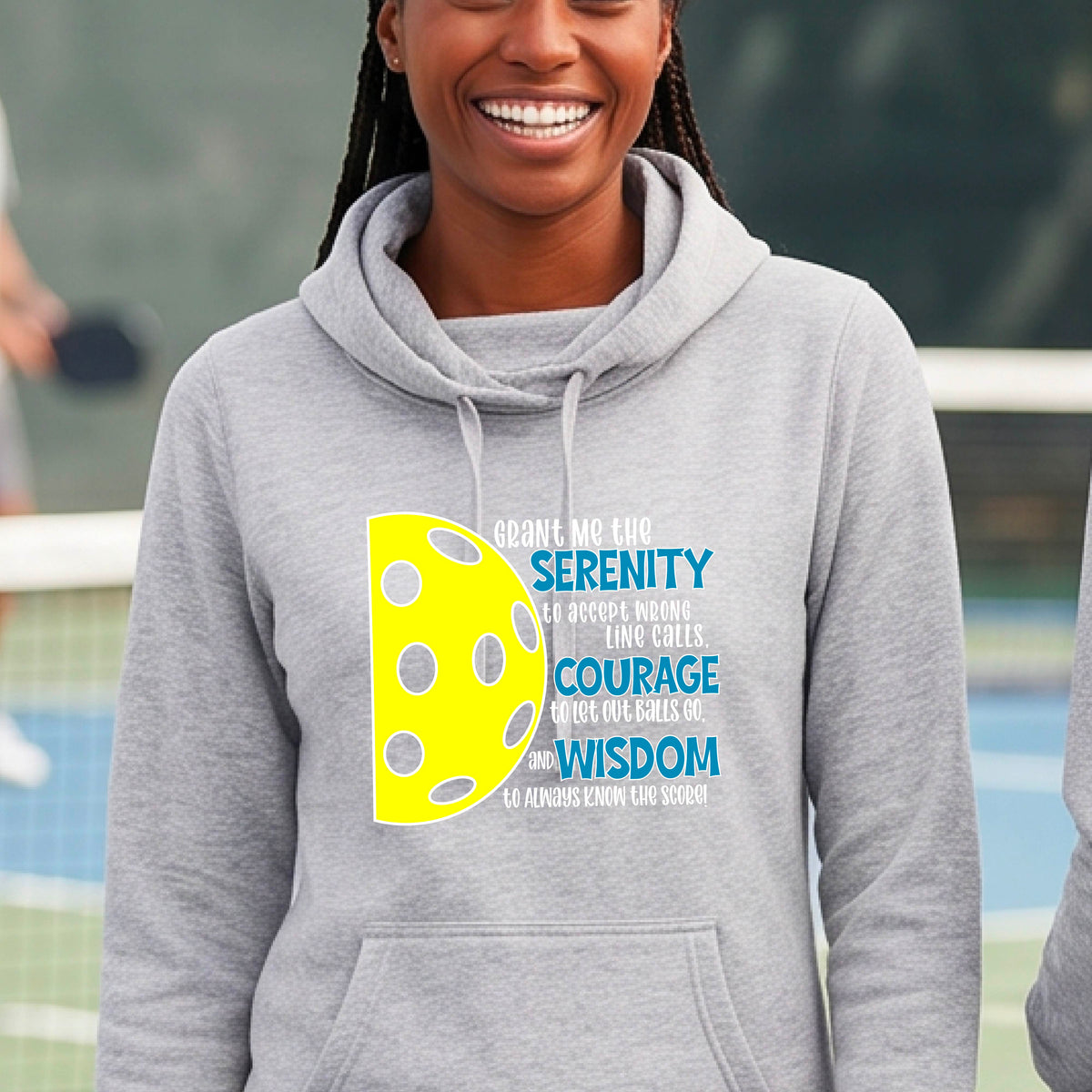 A woman on a pickleball court wearing a gray cowl-neck hoodie pullover with a graphic of the Serenity Prayer made for pickleball:  “Grant me the serenity to Accept Wrong Line Calls; Courage to Let Out Balls Go; and, Wisdom to Always Know the Score” next to a half-pickleball icon, standing on a pickleball court.