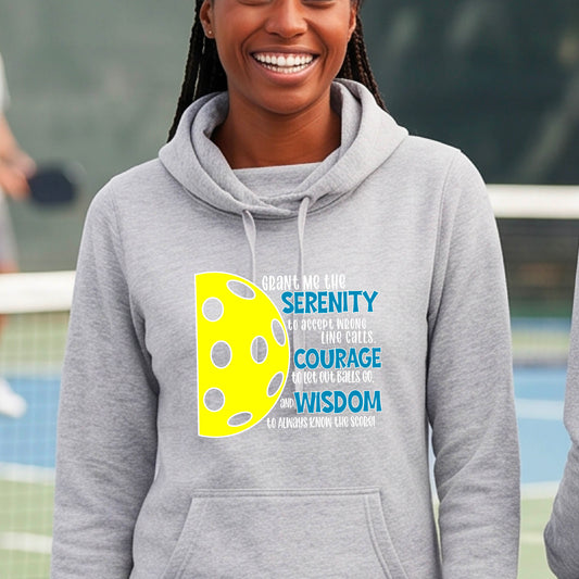 A woman on a pickleball court wearing a gray cowl-neck hoodie pullover with a graphic of the Serenity Prayer made for pickleball:  “Grant me the serenity to Accept Wrong Line Calls; Courage to Let Out Balls Go; and, Wisdom to Always Know the Score” next to a half-pickleball icon, standing on a pickleball court.