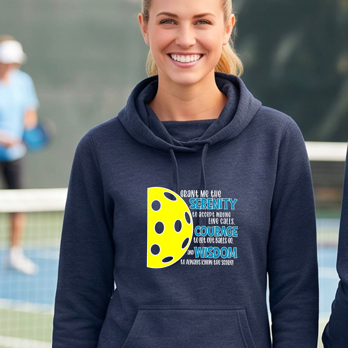 A woman on a pickleball court wearing a navy blue cowl-neck hoodie pullover with a graphic of the Serenity Prayer made for pickleball:  “Grant me the serenity to Accept Wrong Line Calls; Courage to Let Out Balls Go; and, Wisdom to Always Know the Score” next to a half-pickleball icon, standing on a pickleball court.
