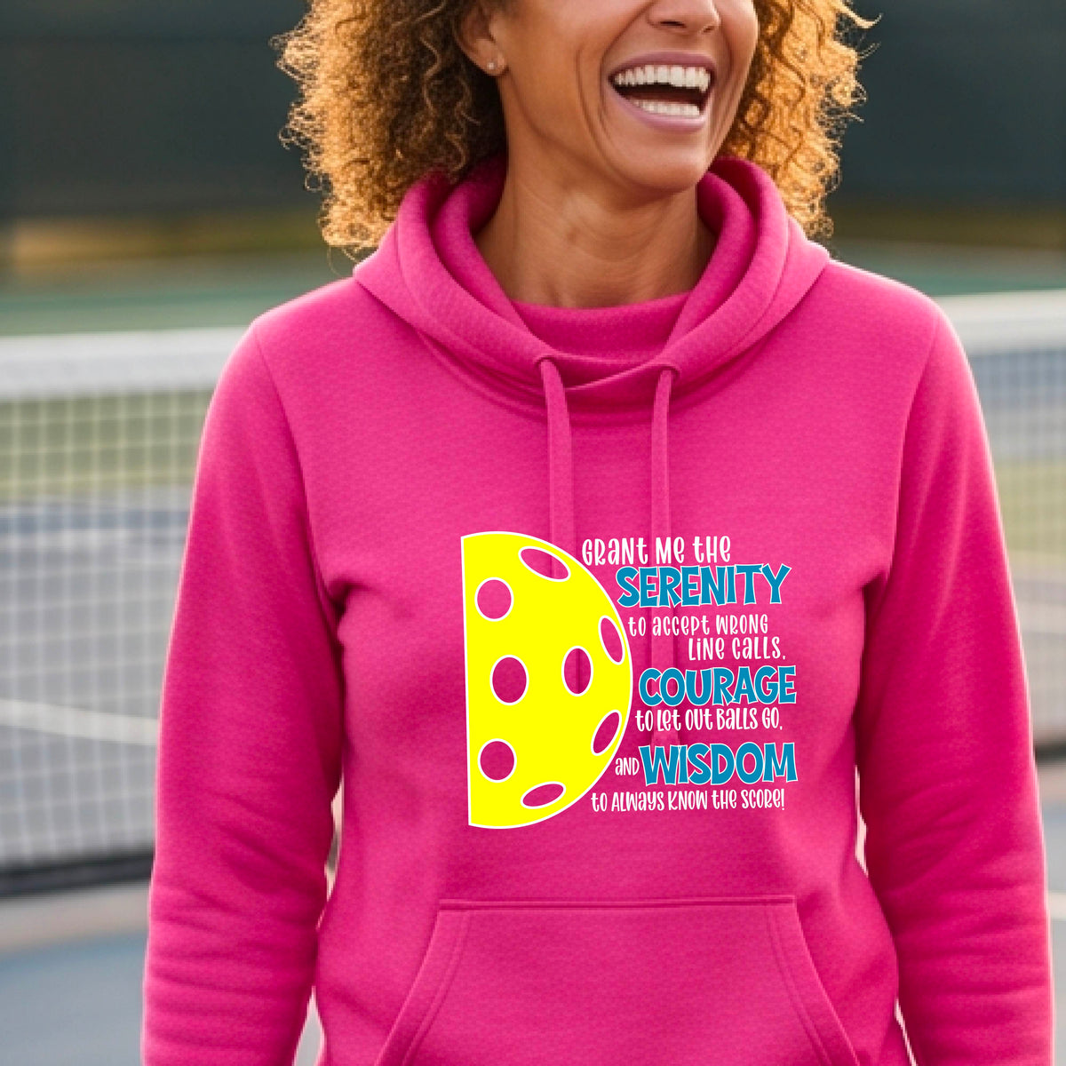 A woman on a pickleball court wearing a pink cowl-neck hoodie pullover with a graphic of the Serenity Prayer made for pickleball:  “Grant me the serenity to Accept Wrong Line Calls; Courage to Let Out Balls Go; and, Wisdom to Always Know the Score” next to a half-pickleball icon, standing on a pickleball court.