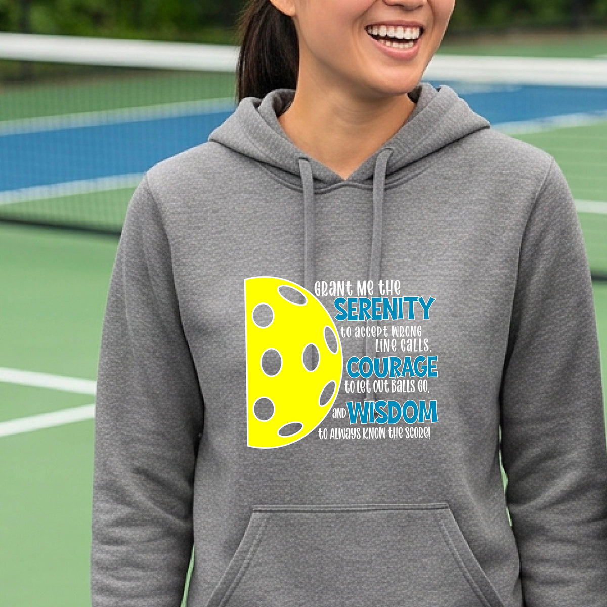 A woman on a pickleball court wearing a gray fitted hoodie pullover with a graphic of the Serenity Prayer made for pickleball:  “Grant me the serenity to Accept Wrong Line Calls; Courage to Let Out Balls Go; and, Wisdom to Always Know the Score” next to a half-pickleball icon, standing on a pickleball court.