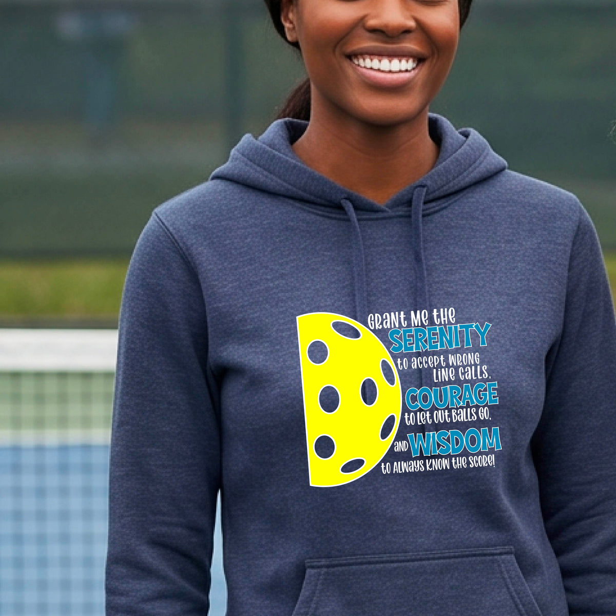 A woman on a pickleball court wearing a navy blue fitted hoodie pullover with a graphic of the Serenity Prayer made for pickleball:  “Grant me the serenity to Accept Wrong Line Calls; Courage to Let Out Balls Go; and, Wisdom to Always Know the Score” next to a half-pickleball icon, standing on a pickleball court.