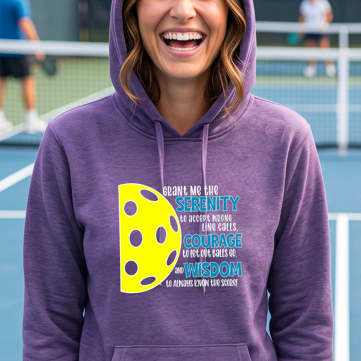 A woman on a pickleball court wearing a purple fitted hoodie pullover with a graphic of the Serenity Prayer made for pickleball:  “Grant me the serenity to Accept Wrong Line Calls; Courage to Let Out Balls Go; and, Wisdom to Always Know the Score” next to a half-pickleball icon, standing on a pickleball court.