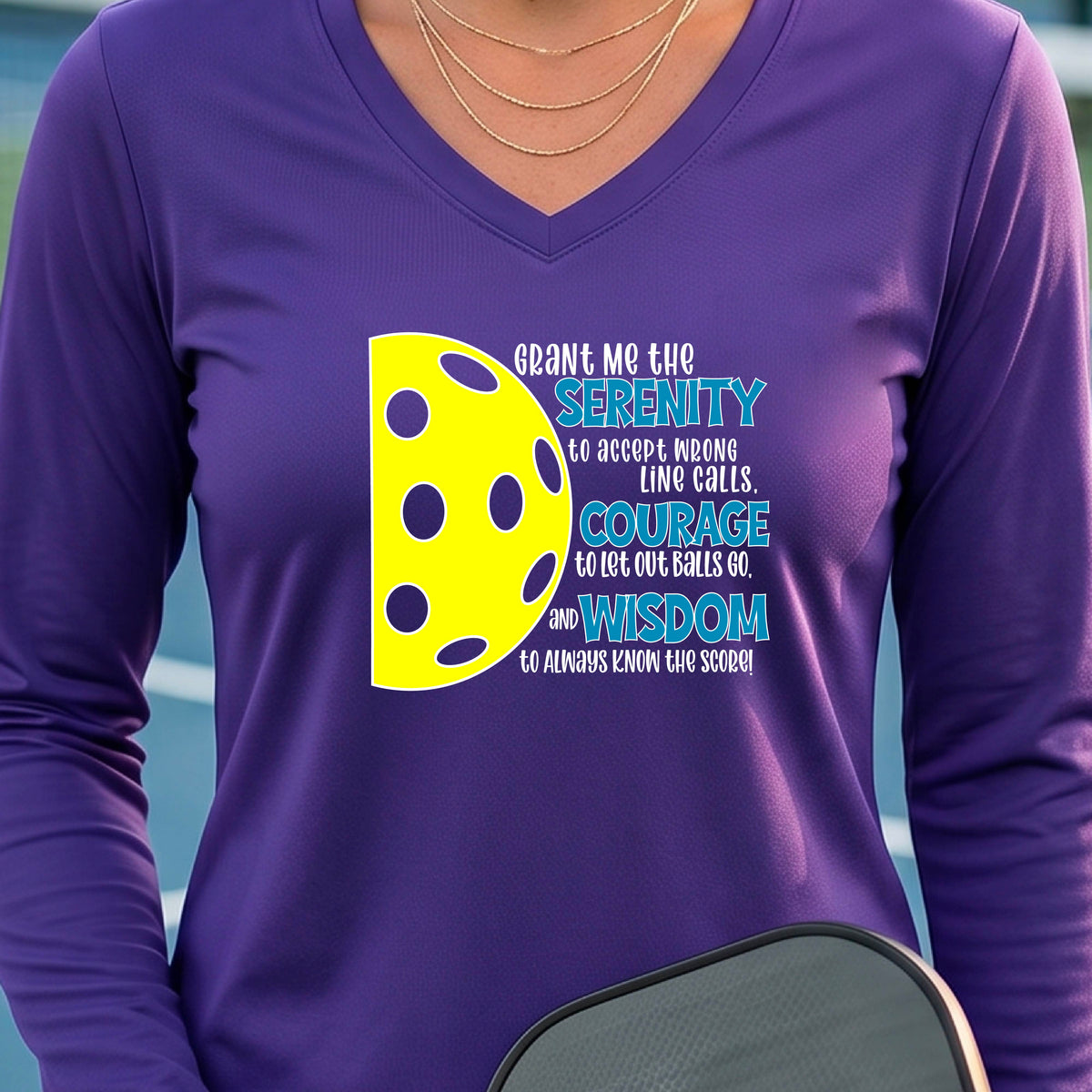 A woman on a pickleball court wearing a purple long-sleeve t-shirt with a graphic of the Serenity Prayer made for pickleball:  “Grant me the serenity to Accept Wrong Line Calls; Courage to Let Out Balls Go; and, Wisdom to Always Know the Score” next to a half-pickleball icon, standing on a pickleball court.