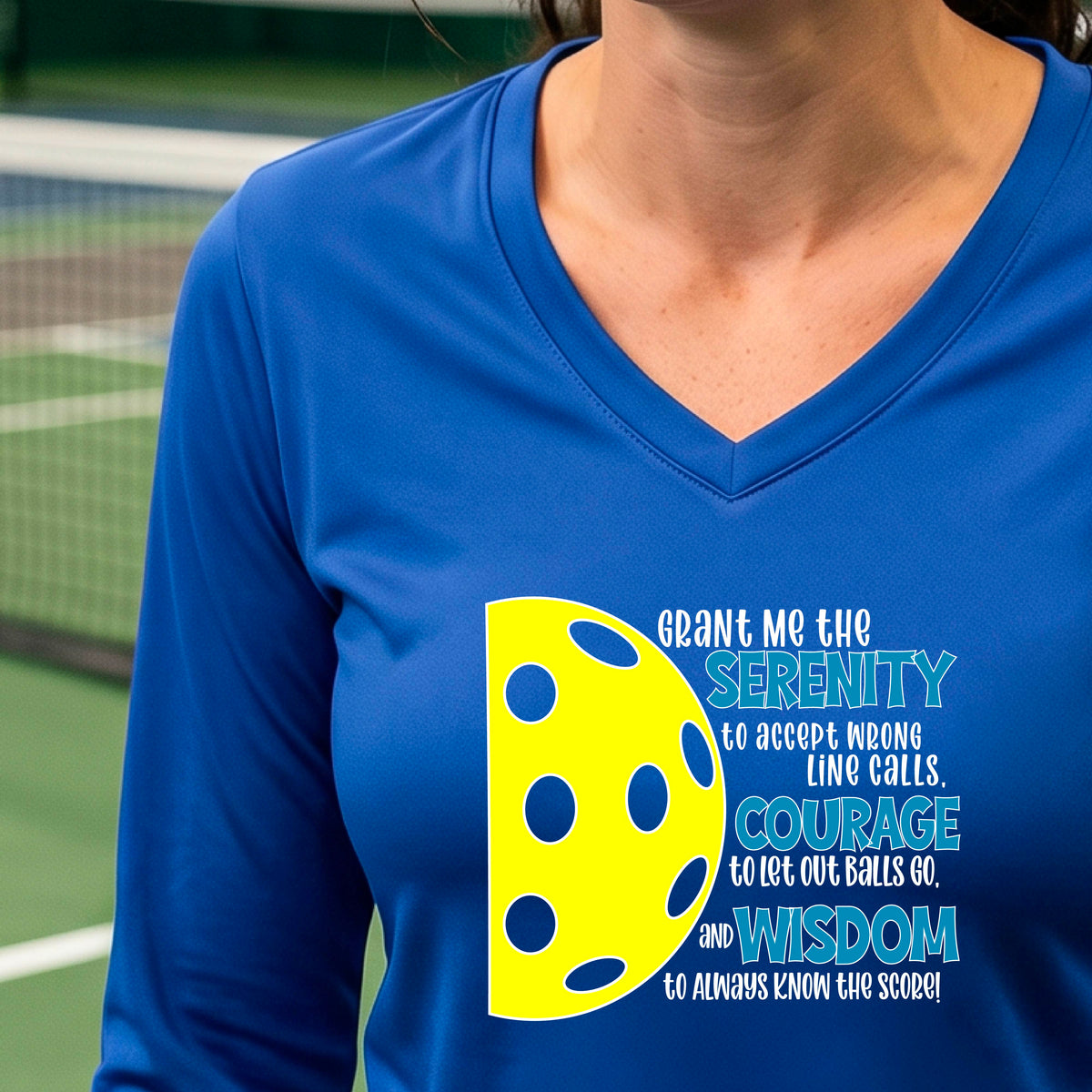 A woman on a pickleball court wearing a royal blue long-sleeve t-shirt with a graphic of the Serenity Prayer made for pickleball:  “Grant me the serenity to Accept Wrong Line Calls; Courage to Let Out Balls Go; and, Wisdom to Always Know the Score” next to a half-pickleball icon, standing on a pickleball court.