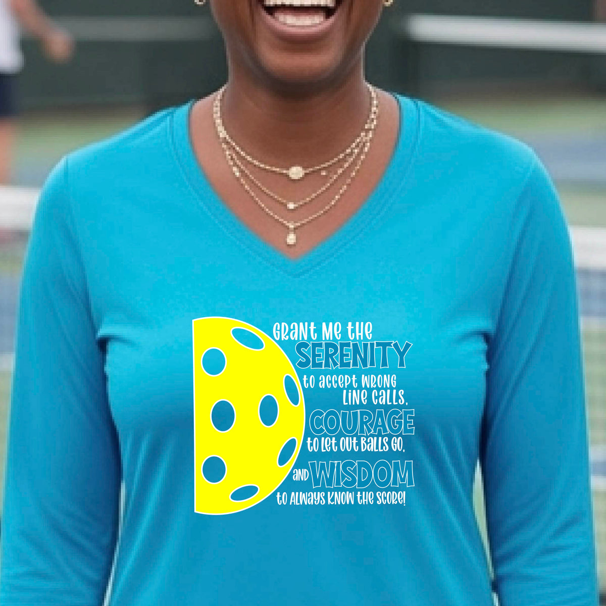 A woman on a pickleball court wearing a bright blue long-sleeve t-shirt with a graphic of the Serenity Prayer made for pickleball:  “Grant me the serenity to Accept Wrong Line Calls; Courage to Let Out Balls Go; and, Wisdom to Always Know the Score” next to a half-pickleball icon, standing on a pickleball court.