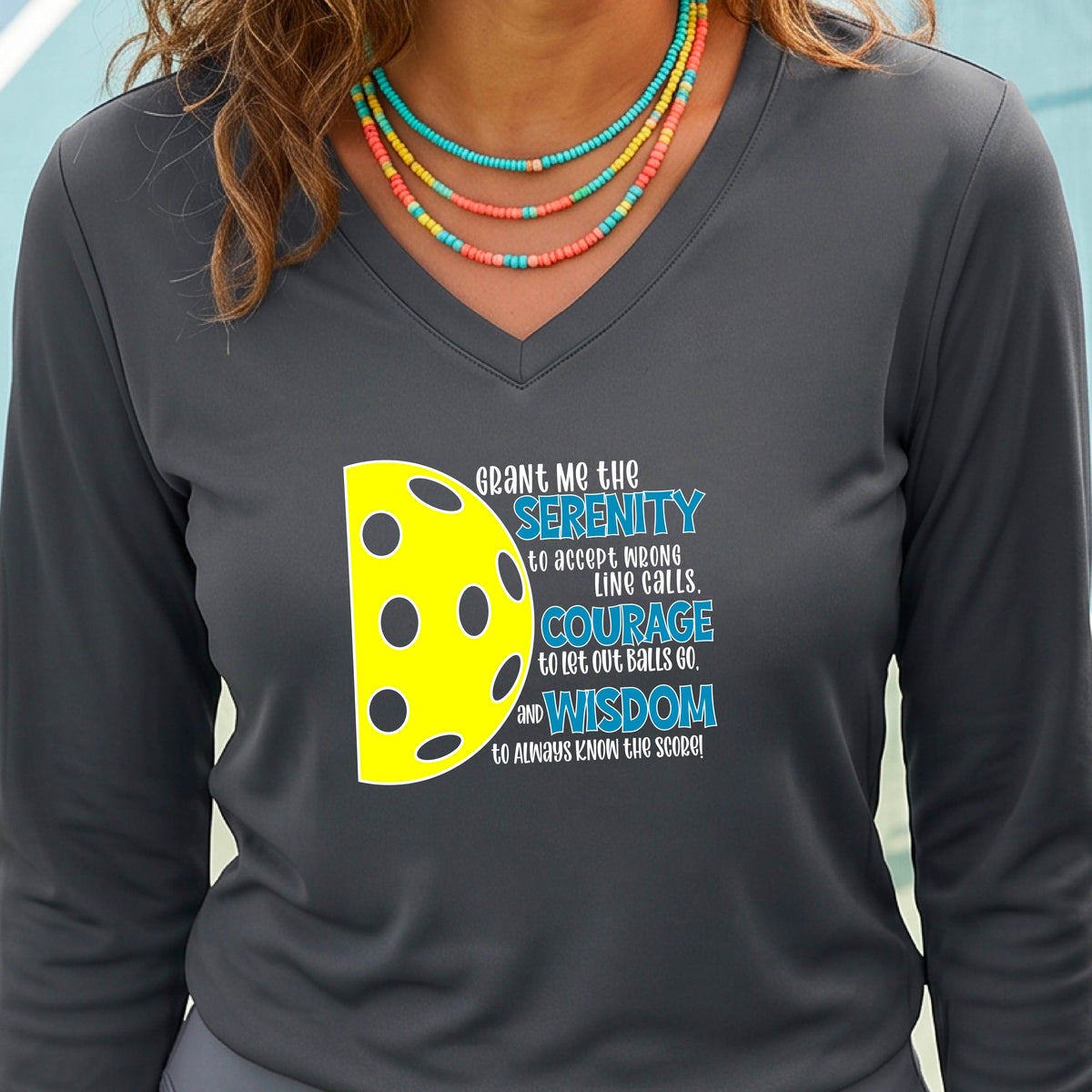A woman on a pickleball court wearing a dark gray long-sleeve t-shirt with a graphic of the Serenity Prayer made for pickleball:  “Grant me the serenity to Accept Wrong Line Calls; Courage to Let Out Balls Go; and, Wisdom to Always Know the Score” next to a half-pickleball icon, standing on a pickleball court.