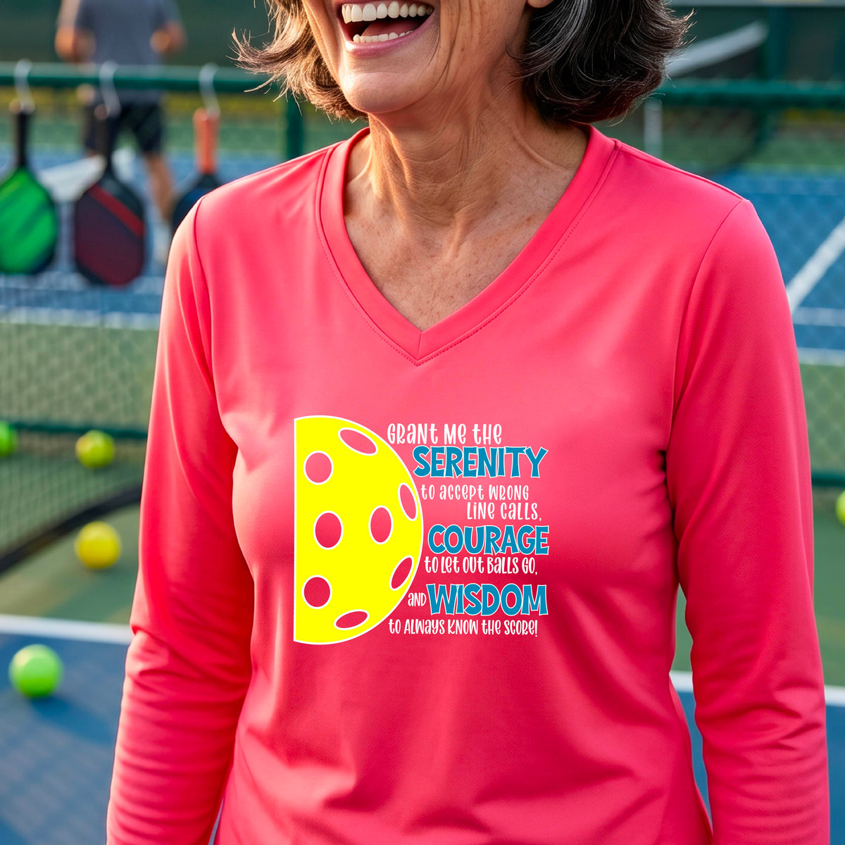 A woman on a pickleball court wearing a bright coral pink long-sleeve t-shirt with a graphic of the Serenity Prayer made for pickleball:  “Grant me the serenity to Accept Wrong Line Calls; Courage to Let Out Balls Go; and, Wisdom to Always Know the Score” next to a half-pickleball icon, standing on a pickleball court.