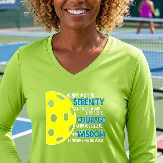 A woman on a pickleball court wearing a lime green long-sleeve t-shirt with a graphic of the Serenity Prayer made for pickleball:  “Grant me the serenity to Accept Wrong Line Calls; Courage to Let Out Balls Go; and, Wisdom to Always Know the Score” next to a half-pickleball icon, standing on a pickleball court.