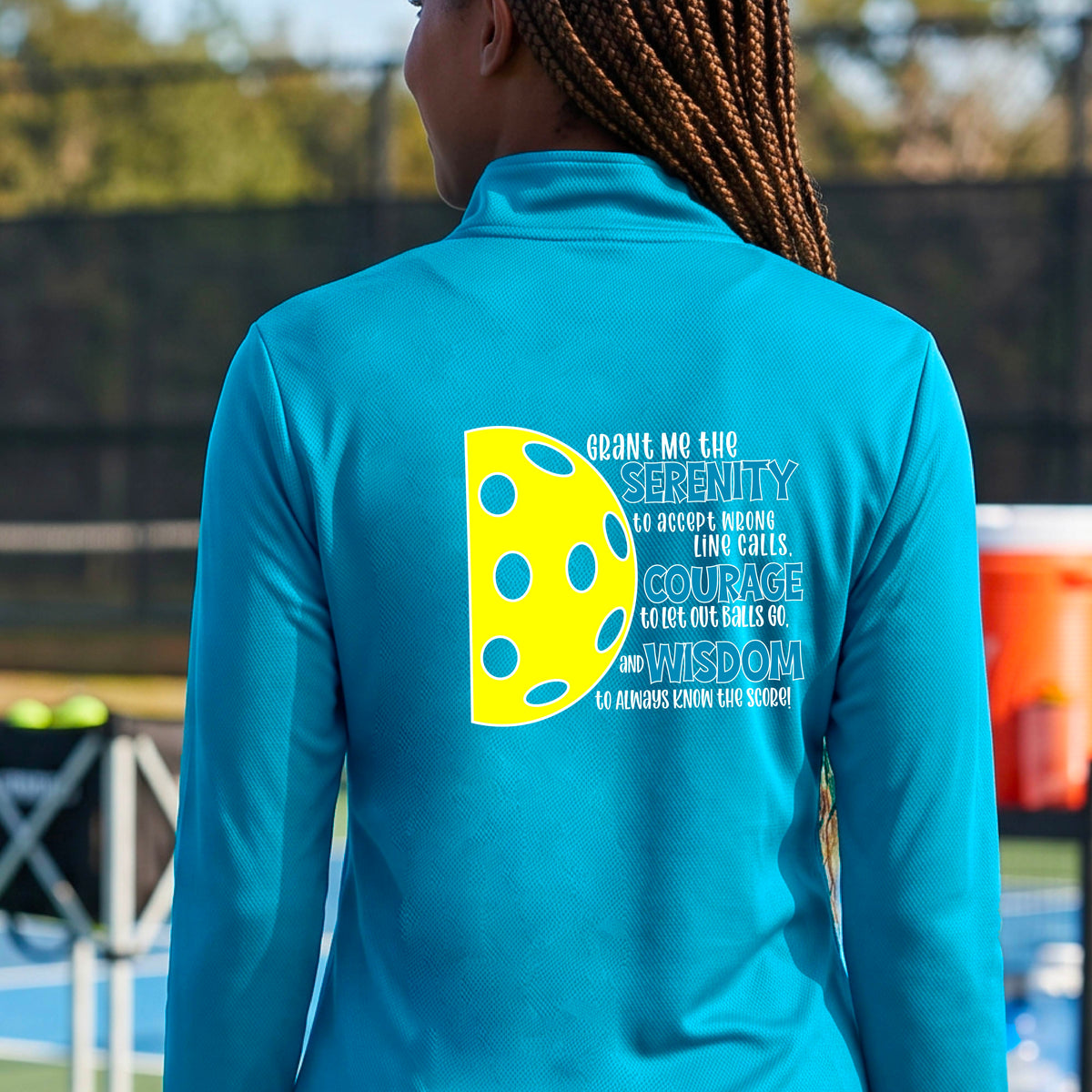 A woman on a pickleball court wearing a bright blue quarter-zip shirt , facing backwards with a graphic of the Serenity Prayer made for pickleball:  “Grant me the serenity to Accept Wrong Line Calls; Courage to Let Out Balls Go; and, Wisdom to Always Know the Score” next to a half-pickleball icon, standing on a pickleball court. 