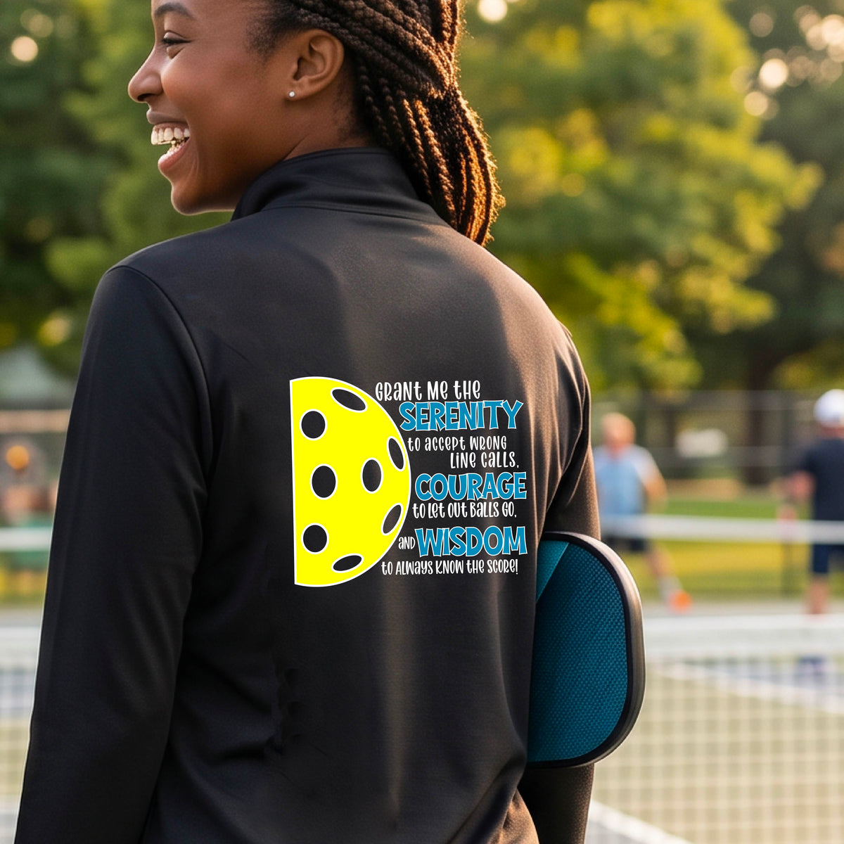 A woman on a pickleball court wearing a black quarter-zip shirt , facing backwards with a graphic of the Serenity Prayer made for pickleball:  “Grant me the serenity to Accept Wrong Line Calls; Courage to Let Out Balls Go; and, Wisdom to Always Know the Score” next to a half-pickleball icon, standing on a pickleball court. 