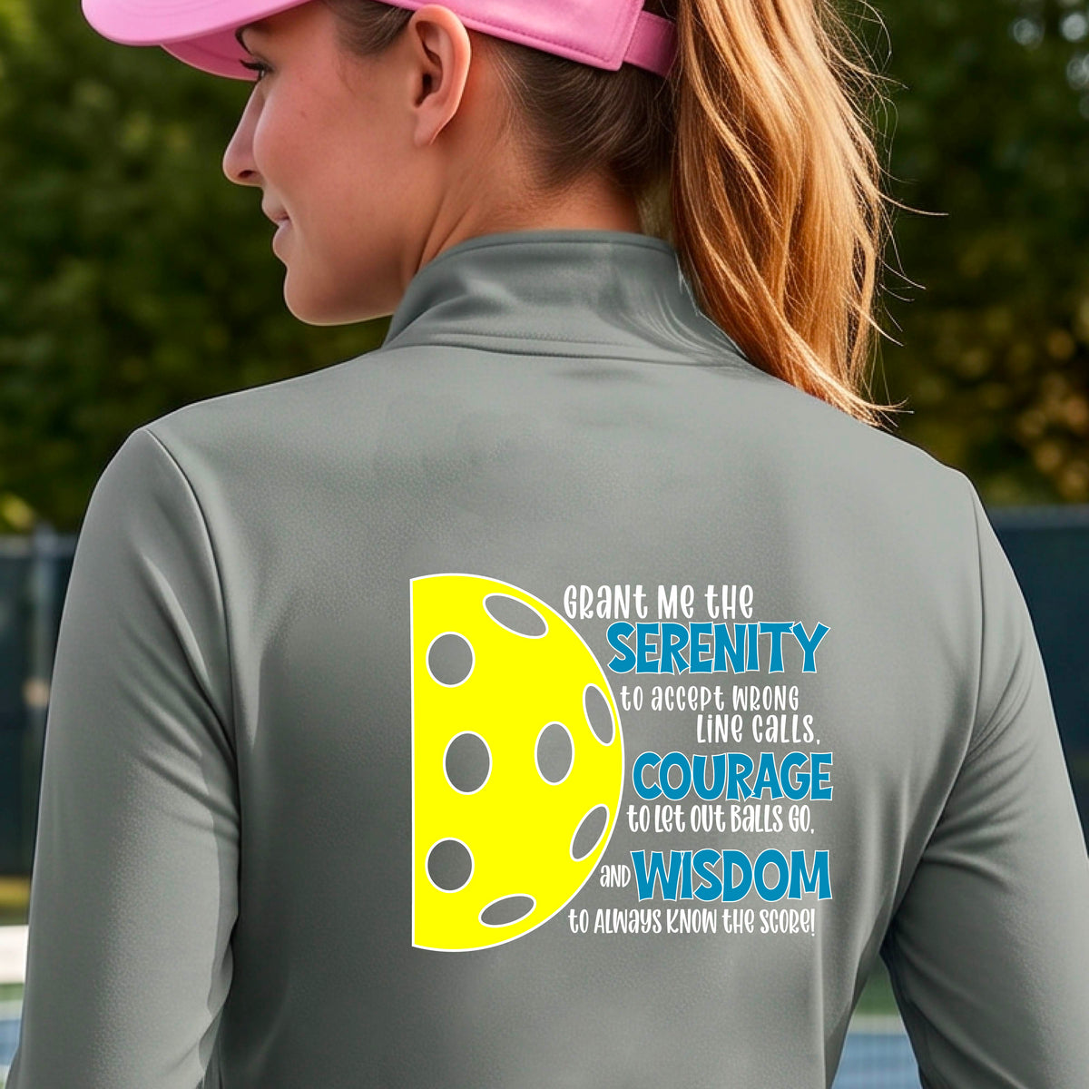 A woman on a pickleball court wearing a gray Quarter-zip shirt , facing backwards with a graphic of the Serenity Prayer made for pickleball:  “Grant me the serenity to Accept Wrong Line Calls; Courage to Let Out Balls Go; and, Wisdom to Always Know the Score” next to a half-pickleball icon, standing on a pickleball court. 