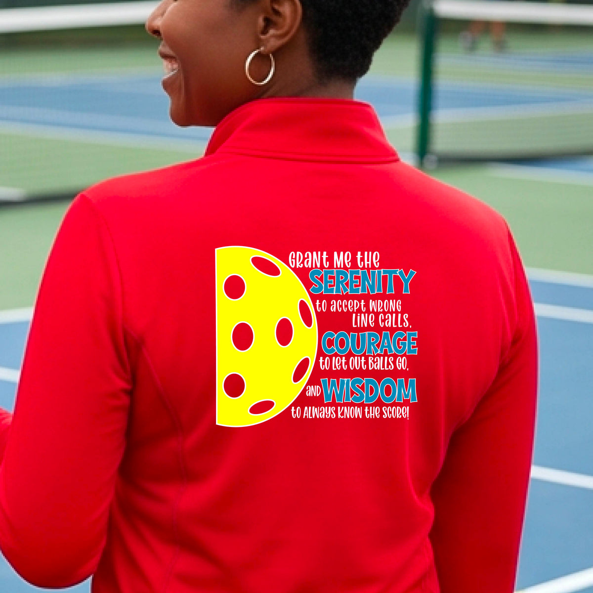 A woman on a pickleball court wearing a red quarter-zip shirt , facing backwards with a graphic of the Serenity Prayer made for pickleball:  “Grant me the serenity to Accept Wrong Line Calls; Courage to Let Out Balls Go; and, Wisdom to Always Know the Score” next to a half-pickleball icon, standing on a pickleball court. 