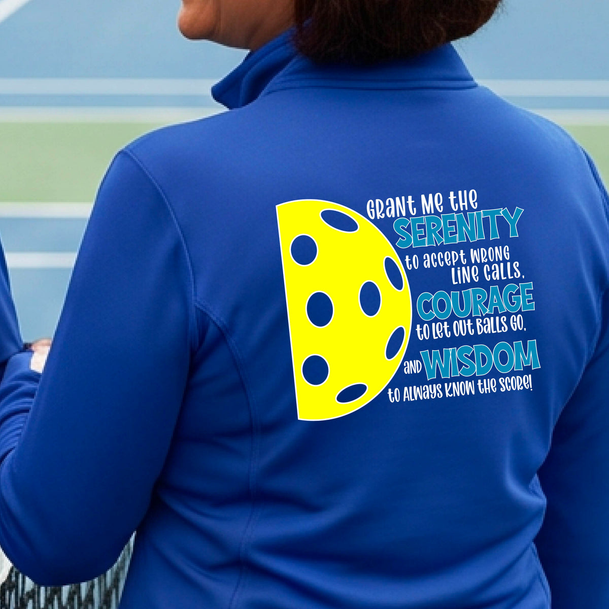 A woman on a pickleball court wearing a royal blue quarter-zip shirt , facing backwards with a graphic of the Serenity Prayer made for pickleball:  “Grant me the serenity to Accept Wrong Line Calls; Courage to Let Out Balls Go; and, Wisdom to Always Know the Score” next to a half-pickleball icon, standing on a pickleball court. 