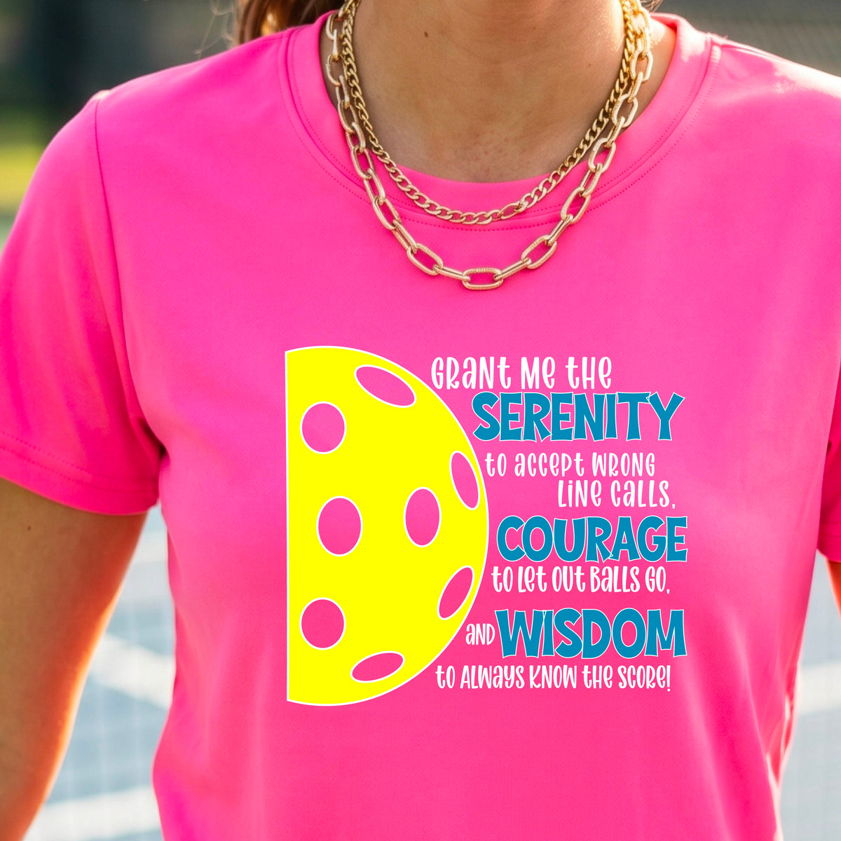 A woman on a pickleball court wearing a pink short-sleeve t-shirt with a graphic of the Serenity Prayer made for pickleball:  “Grant me the serenity to Accept Wrong Line Calls; Courage to Let Out Balls Go; and, Wisdom to Always Know the Score” next to a half-pickleball icon, standing on a pickleball court.