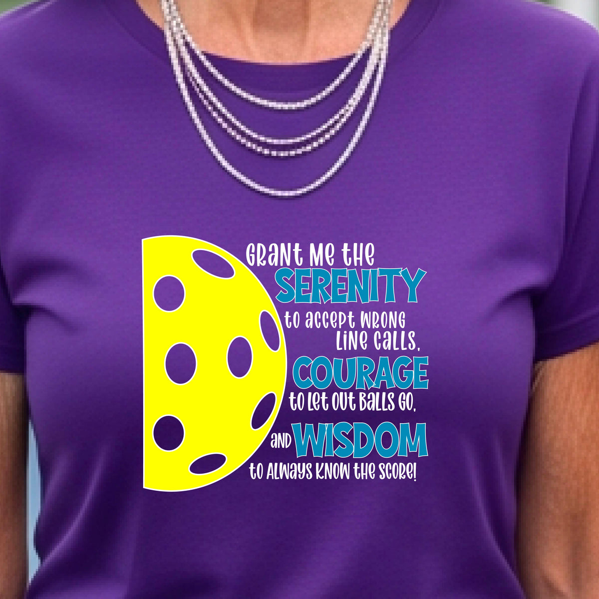 A woman on a pickleball court wearing a purple short-sleeve t-shirt with a graphic of the Serenity Prayer made for pickleball:  “Grant me the serenity to Accept Wrong Line Calls; Courage to Let Out Balls Go; and, Wisdom to Always Know the Score” next to a half-pickleball icon, standing on a pickleball court.