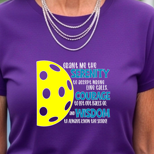 A woman on a pickleball court wearing a purple short-sleeve t-shirt with a graphic of the Serenity Prayer made for pickleball:  “Grant me the serenity to Accept Wrong Line Calls; Courage to Let Out Balls Go; and, Wisdom to Always Know the Score” next to a half-pickleball icon, standing on a pickleball court.