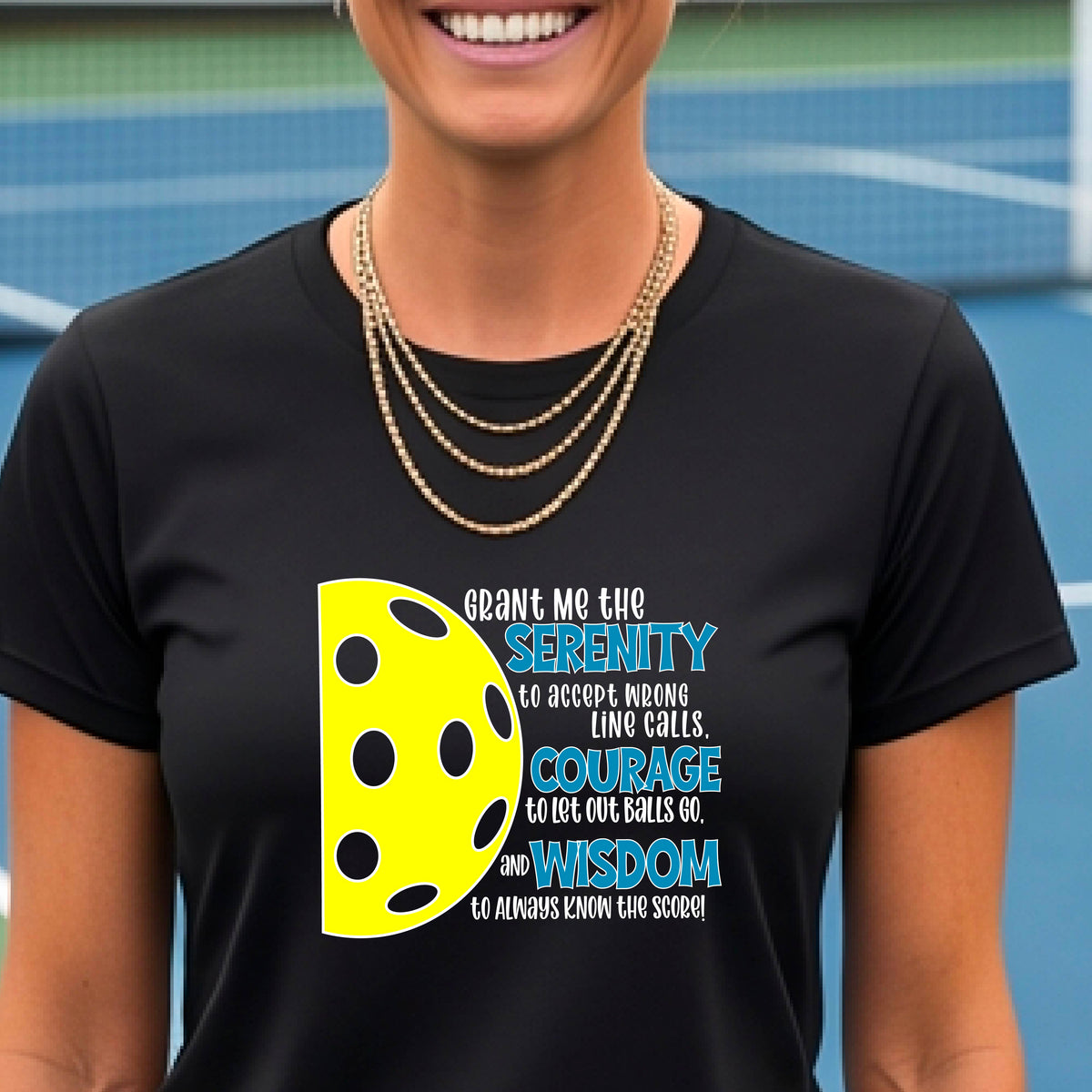 A woman on a pickleball court wearing a black short-sleeve t-shirt with a graphic of the Serenity Prayer made for pickleball:  “Grant me the serenity to Accept Wrong Line Calls; Courage to Let Out Balls Go; and, Wisdom to Always Know the Score” next to a half-pickleball icon, standing on a pickleball court.