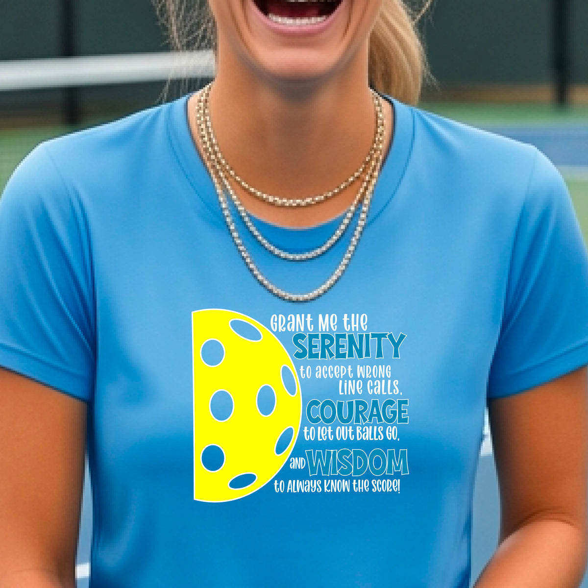 A woman on a pickleball court wearing a blue short-sleeve t-shirt with a graphic of the Serenity Prayer made for pickleball:  “Grant me the serenity to Accept Wrong Line Calls; Courage to Let Out Balls Go; and, Wisdom to Always Know the Score” next to a half-pickleball icon, standing on a pickleball court.