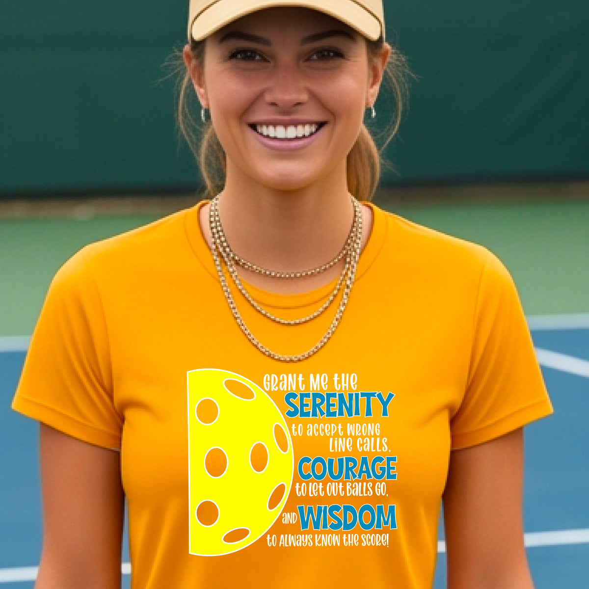 A woman on a pickleball court wearing a mustard yellow gold short-sleeve t-shirt with a graphic of the Serenity Prayer made for pickleball:  “Grant me the serenity to Accept Wrong Line Calls; Courage to Let Out Balls Go; and, Wisdom to Always Know the Score” next to a half-pickleball icon, standing on a pickleball court.